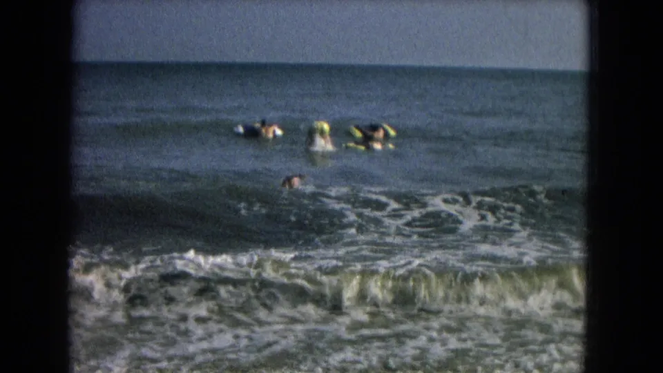 1967 8mm frame showing swimmers and bodyboarders near the shore break, with white foam, open water, and horizon behind them.