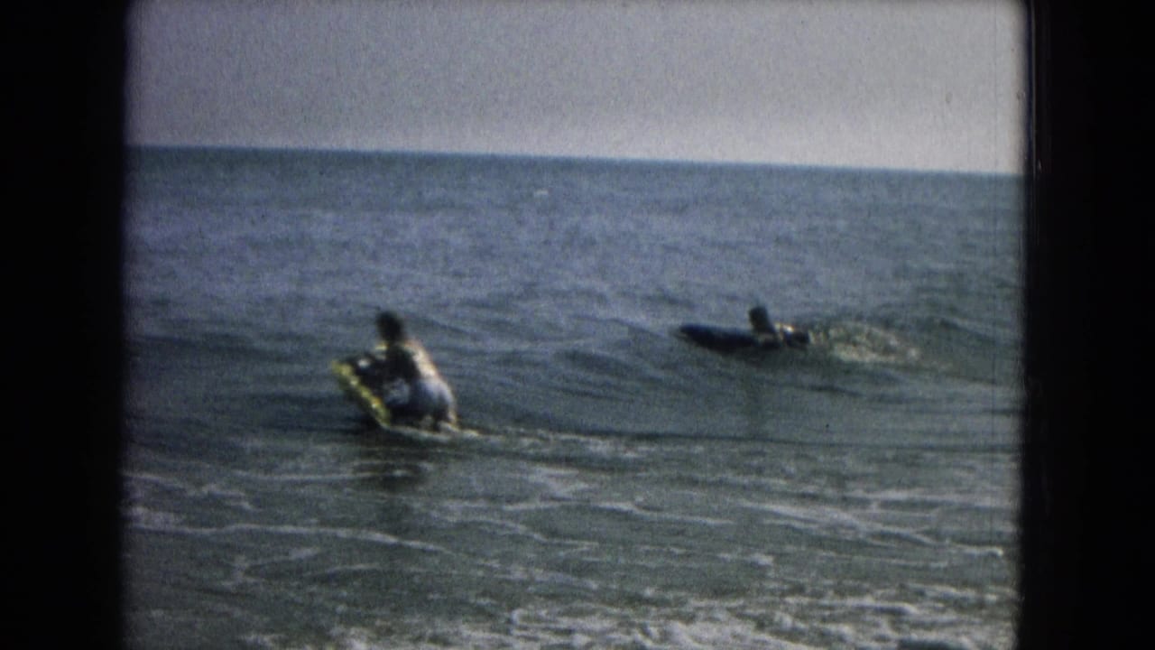 Home-movie frame showing two distant people on boards or flotation gear beyond gentle surf.