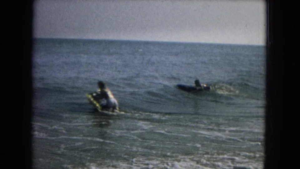 Home-movie frame showing two distant people on boards or flotation gear beyond gentle surf.