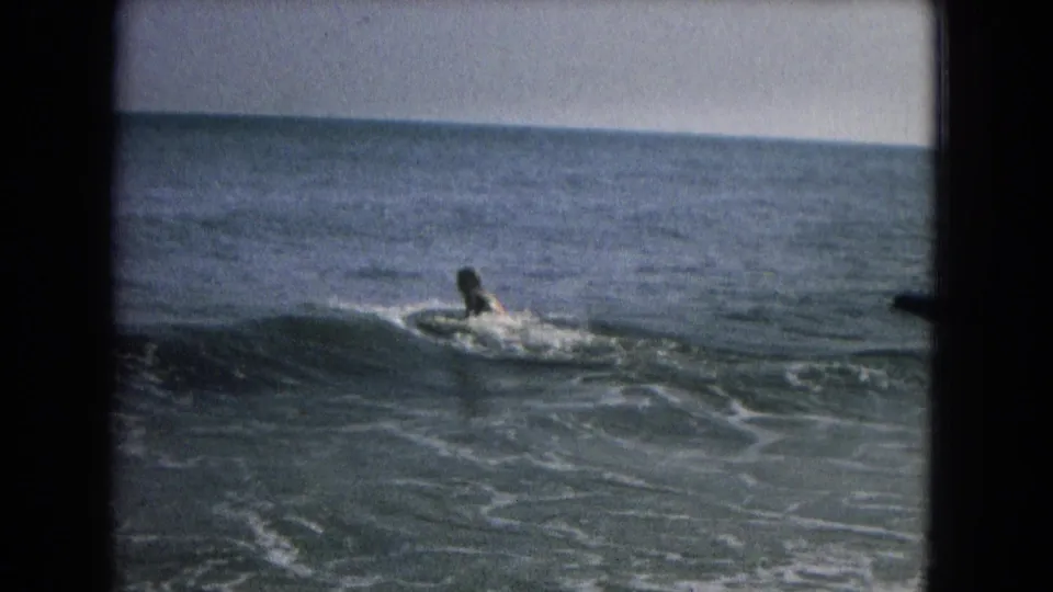 A distant swimmer or surfer paddles on a board in choppy water, filmed in soft 8mm color.