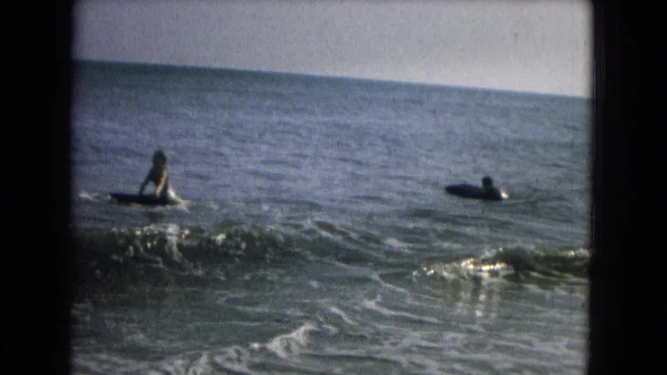 1960s home-movie frame of two distant board riders in gentle surf with foam crossing the foreground.