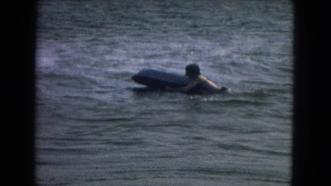 A person appears in calm water beside a thick board, with soft 8mm grain and dark contact-sheet borders around the frame.