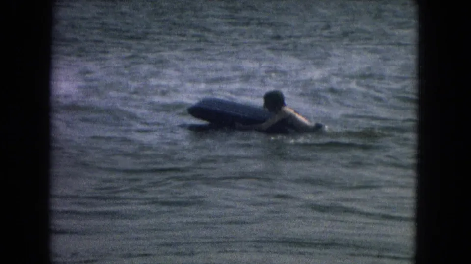 A person appears in calm water beside a thick board, with soft 8mm grain and dark contact-sheet borders around the frame.