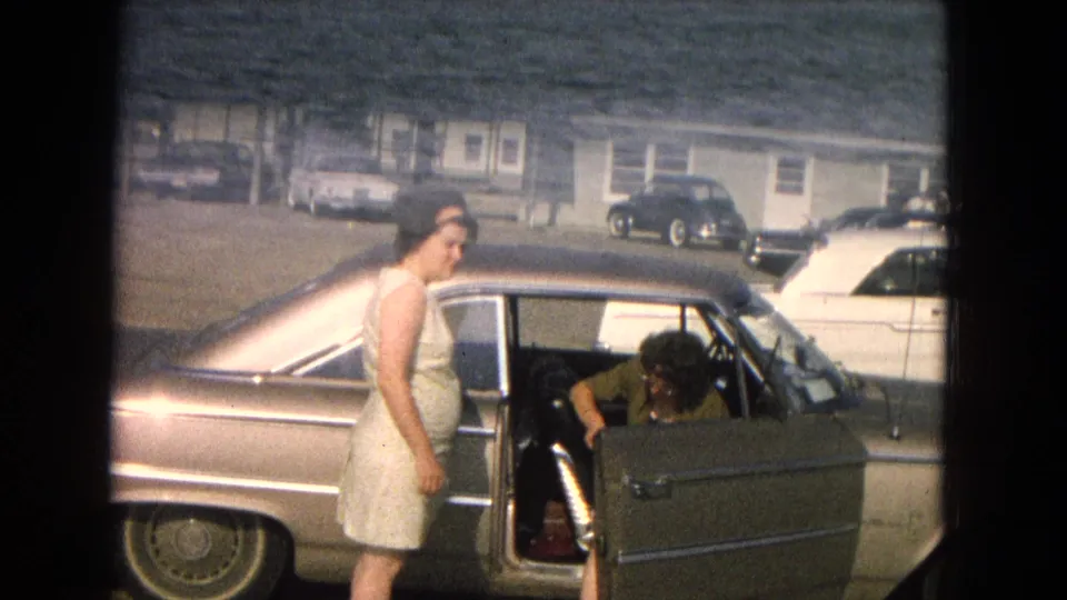 A woman in a sleeveless dress stands by an open mid-century car door, with another person inside and parked cars near a low roadside building.