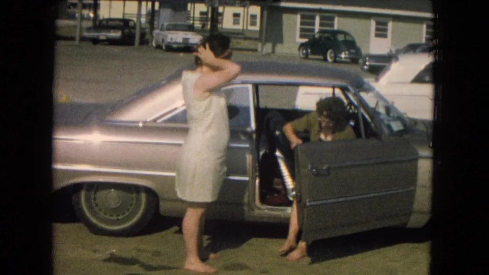 Late-1960s home-movie view of a woman near a parked sedan as a seated person leans at the open door.