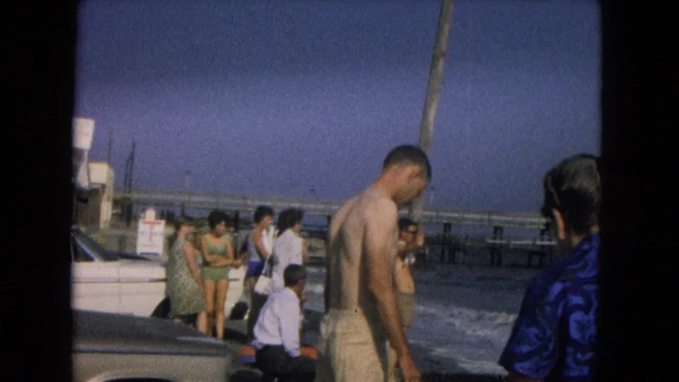 1967 New Mexico lakeside frame showing adults gathered near the water, with a shirtless man in swim trunks in front.