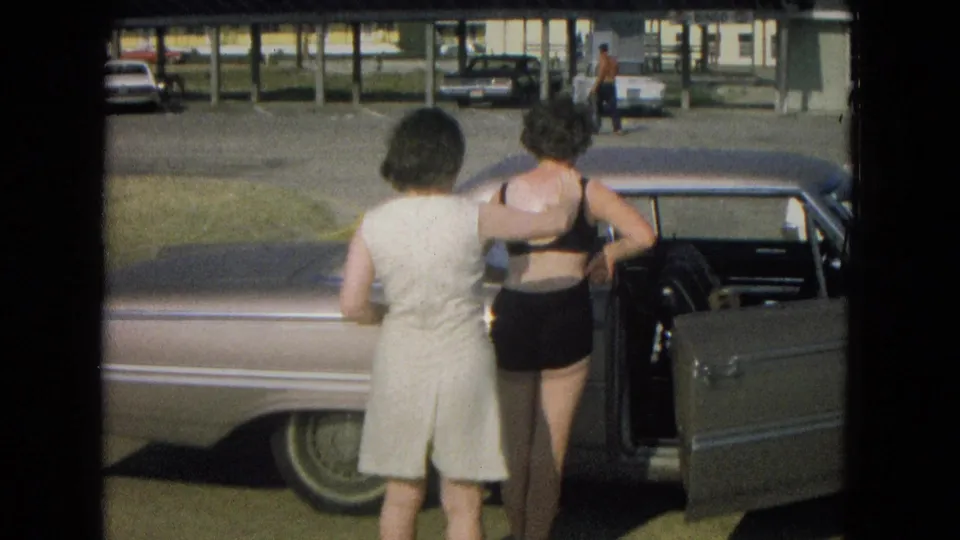 In 1967 New Mexico home-movie footage, two adult women stand by an open door of a light-colored sedan near other parked cars.