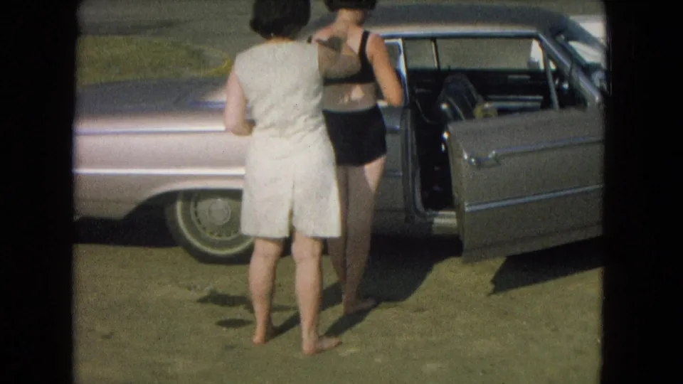 Two adult women, one in a light outfit and one in a dark bathing suit, stand beside an open sedan door in bright outdoor light.