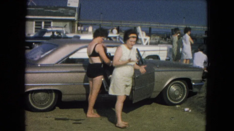 At a New Mexico parking area, one woman walks past the camera while another stands at the open door of a mid-century sedan.