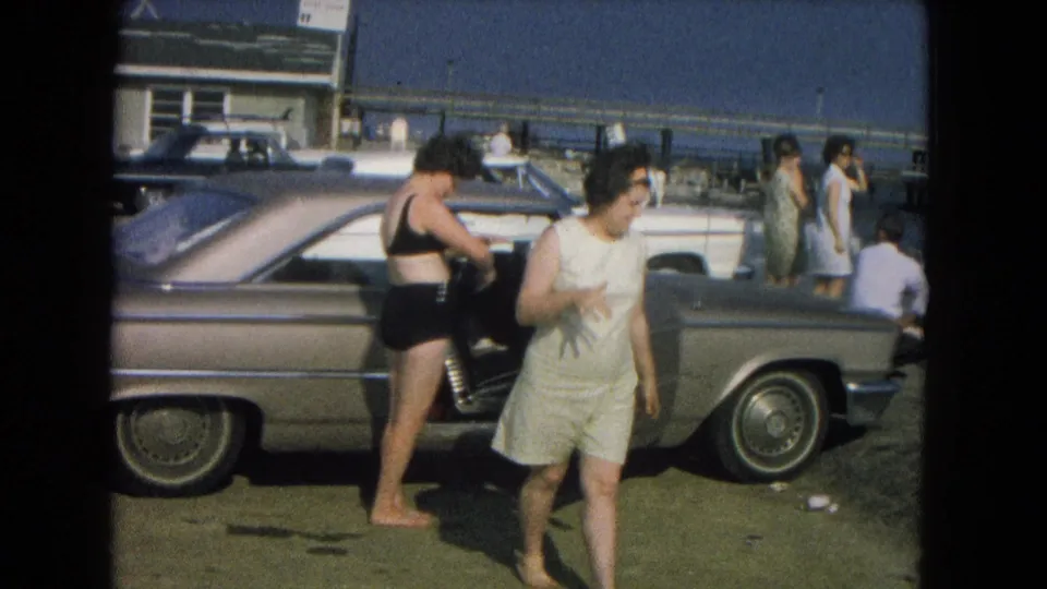The frame shows a woman moving across the foreground and another by the open car door, with people and vehicles behind them.