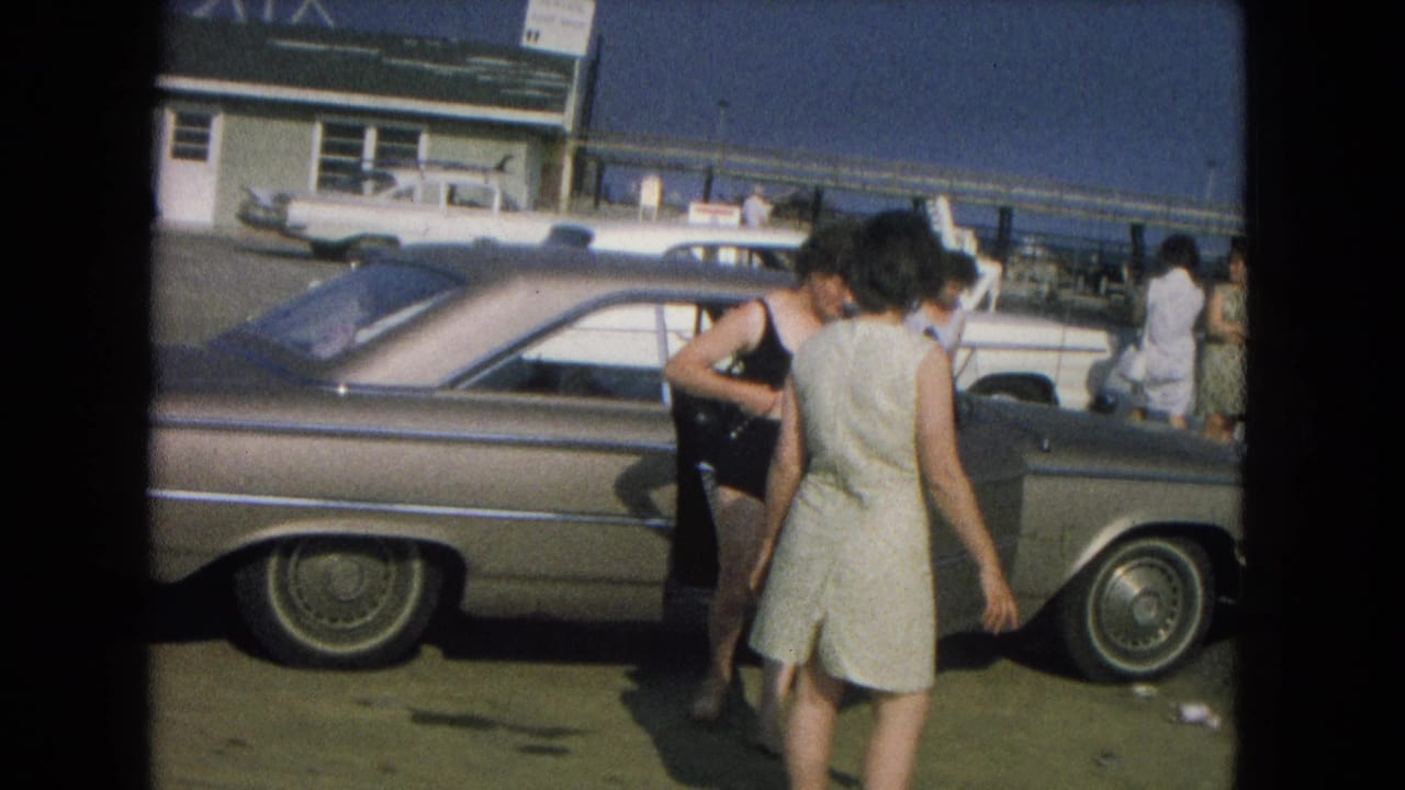 Two women beside a parked 1960s sedan near ocean waves and a pier.
