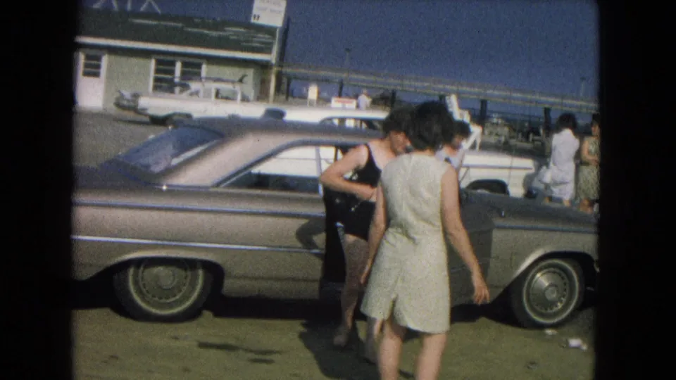 1967 8mm beach scene with a woman in a black bikini near a parked sedan and another woman in a light dress nearby.