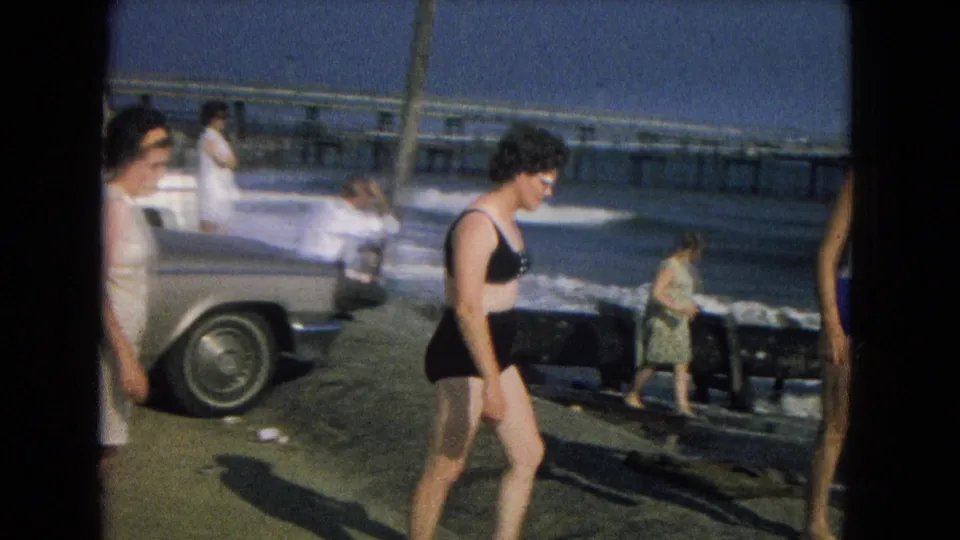 1967 8mm beach footage frame with the bikini-clad woman walking near a seawall, parked car, waves, and other beachgoers.