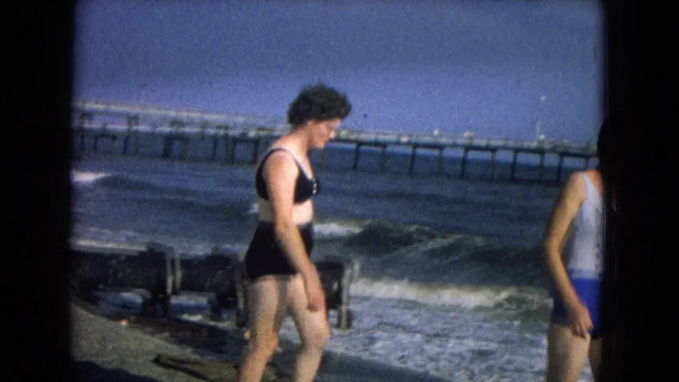 1967 8mm frame of a woman in a black bikini walking beside rough surf, with a long pier extending across the background.