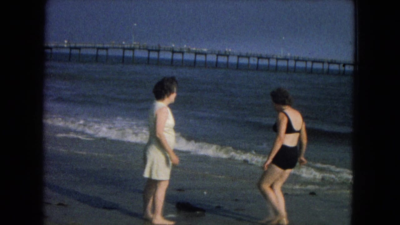 Two women stand barefoot at the shoreline with waves and a long pier behind them.