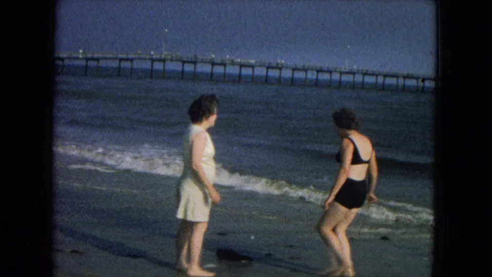In 1967 8mm home-movie footage, two adult women stand near the surf with a long pier visible across the horizon.