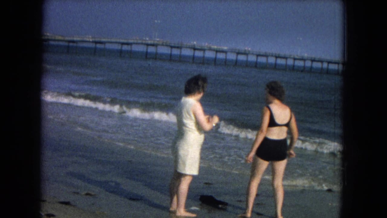 The two adult women stand at the edge of the water, one in a white dress and one in a black swimsuit, with a pier beyond the waves.