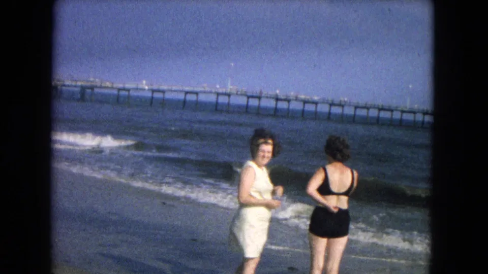 One adult woman turns toward the camera as another faces the surf, with waves and a pier behind them in 1960s 8mm footage.