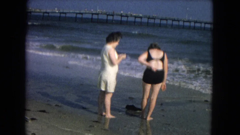 The women remain near the foamy shoreline, one in a white dress and the other bending slightly in a black swimsuit, with the pier in view.