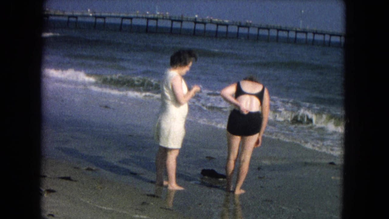 1967 8mm home-movie frame of two women at a beach shoreline, one in a white dress and one in a black swimsuit, with a pier in the background.