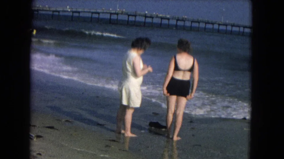 1967 beach home-movie frame showing two women near the waterline, viewed partly from behind, with surf and a long pier beyond them.