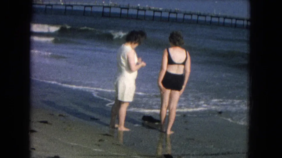 1967 8mm frame of two women paused at the edge of the ocean, with reflective wet sand, waves, and a pier behind them.