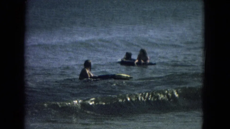 1967 beach home-movie frame showing swimmers in the ocean, with one person close to a surfboard as a small wave approaches.