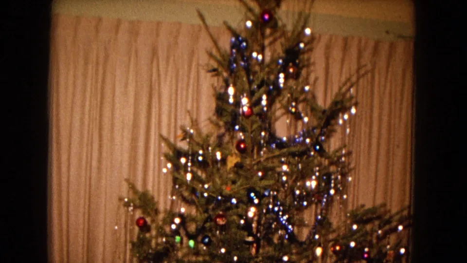 1967 New Mexico home-movie frame showing a lit Christmas tree with ornaments, tinsel, and a star topper before pale curtains.