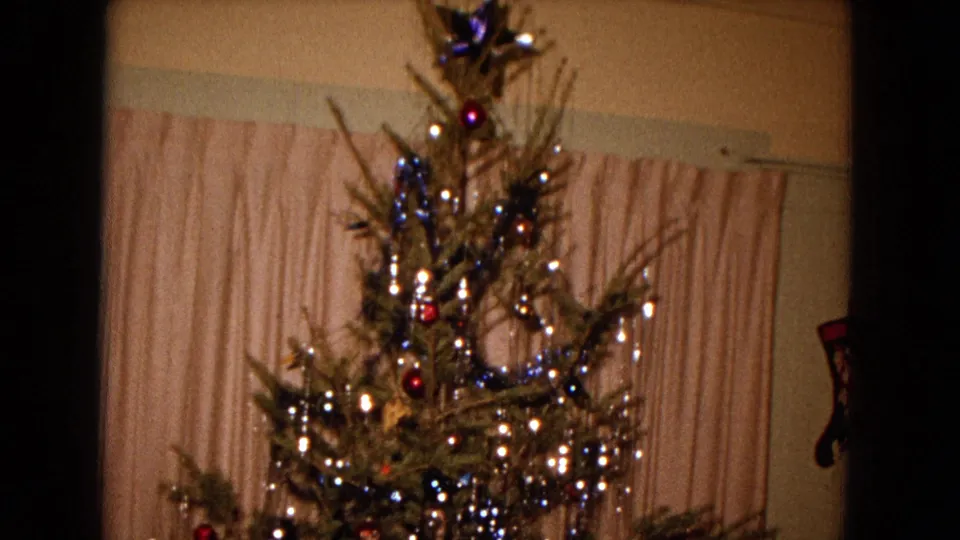 A decorated Christmas tree fills the frame in this 1967 New Mexico home-movie still, with lights, tinsel, ornaments, and a curtain backdrop.
