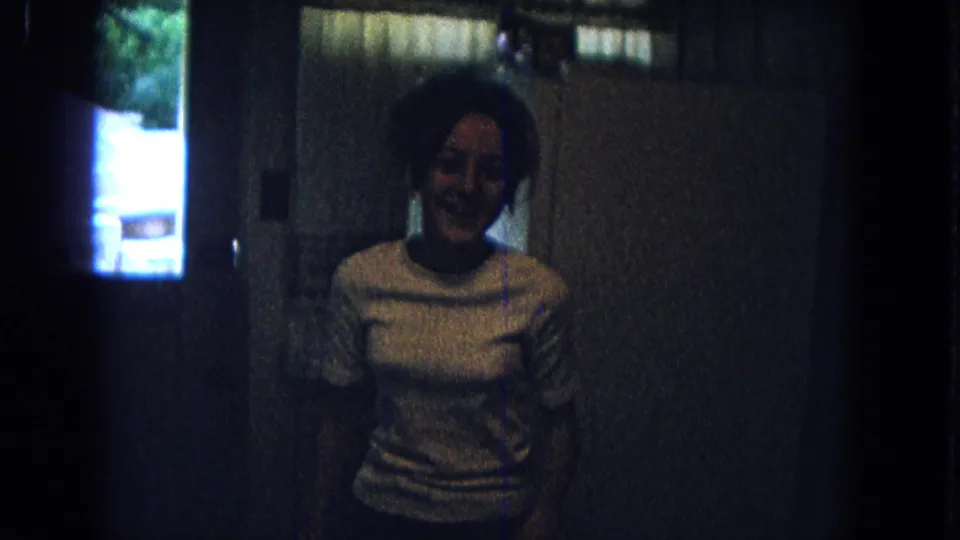 1967 New Mexico home-movie frame showing a young person smiling in a light-colored top inside a dark kitchen, with a window at left.