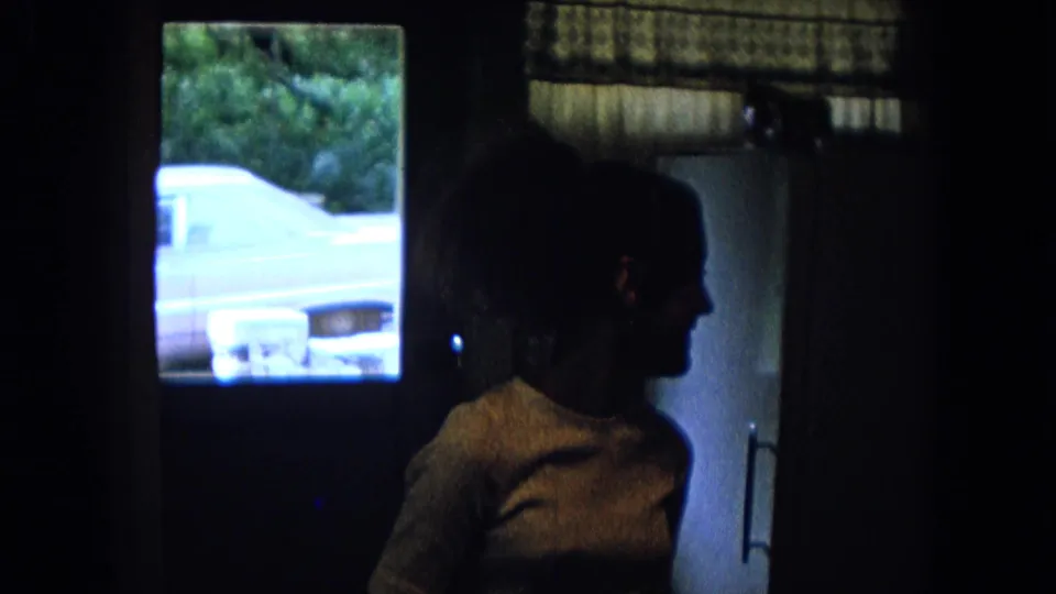 Low-light 8mm frame showing a person in partial profile in a kitchen, with a bright window and vintage car visible outside.