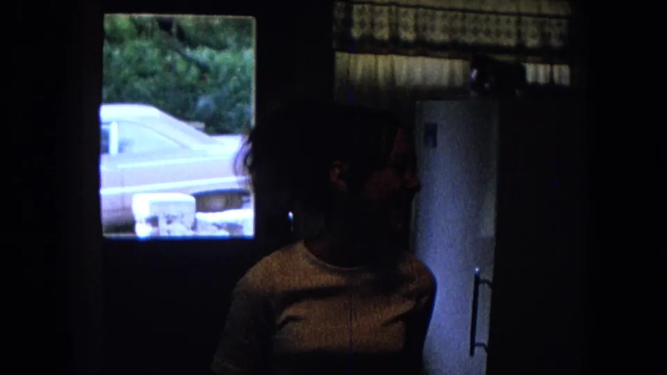 Woman in a dim 1967 New Mexico kitchen, partly silhouetted near a window with a vintage car visible outside.