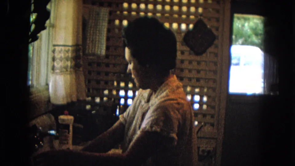Woman at a sink in a 1967 New Mexico kitchen, with a counter bottle, patterned curtains, and bright window light behind her.