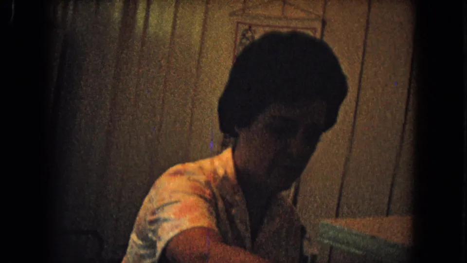 Woman in a light blouse leans near the sink or counter in a dim 1967 New Mexico kitchen with paneled wall behind her.