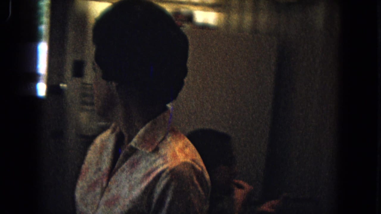 A woman in a floral dress appears in profile inside a dim 1967 New Mexico home kitchen, with a refrigerator behind her.