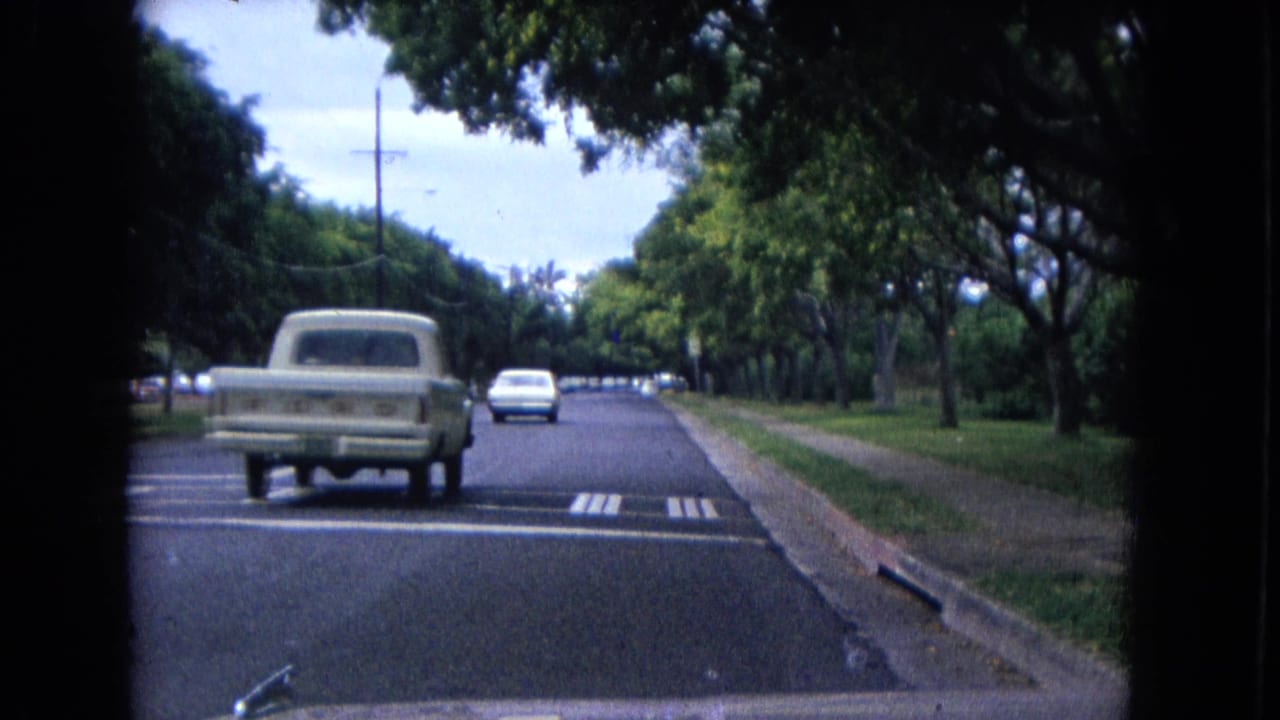 1967 8mm road scene in New Mexico with a light-colored pickup, another car, roadside trees, and the filming vehicle hood in frame.