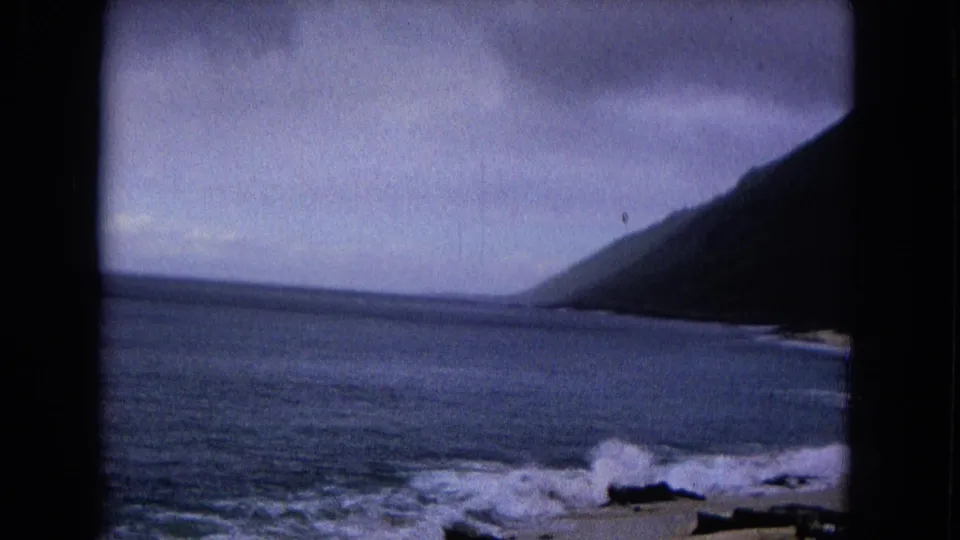 Archival coastal shot with foamy waves, dark rocks, gray sky, and visible 8mm frame borders.