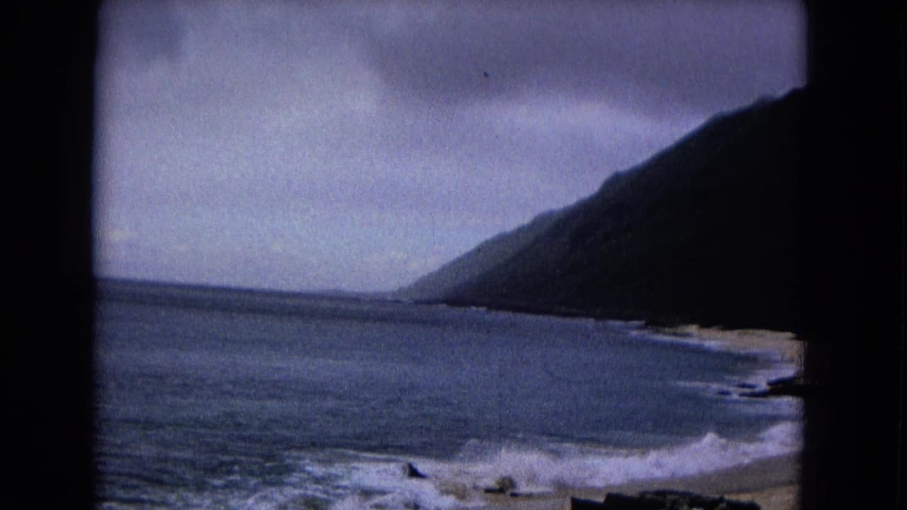 1967 8mm frame showing a steep hillside above a sandy shoreline with surf, cloudy sky, and dark film borders.