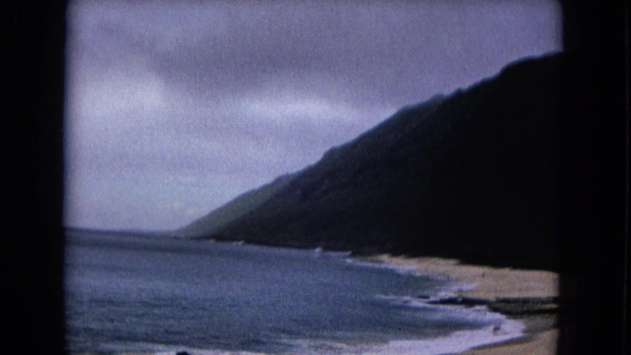 Home-movie frame from 1967 showing a dark coastal slope, pale sand, surf lines, cloudy sky, and 8mm gate edges.
