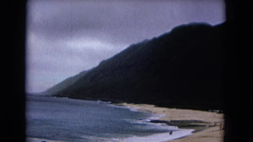 1967 8mm view from above the coast, with a dark hillside, beach, breaking waves, and small distant people near the shoreline.
