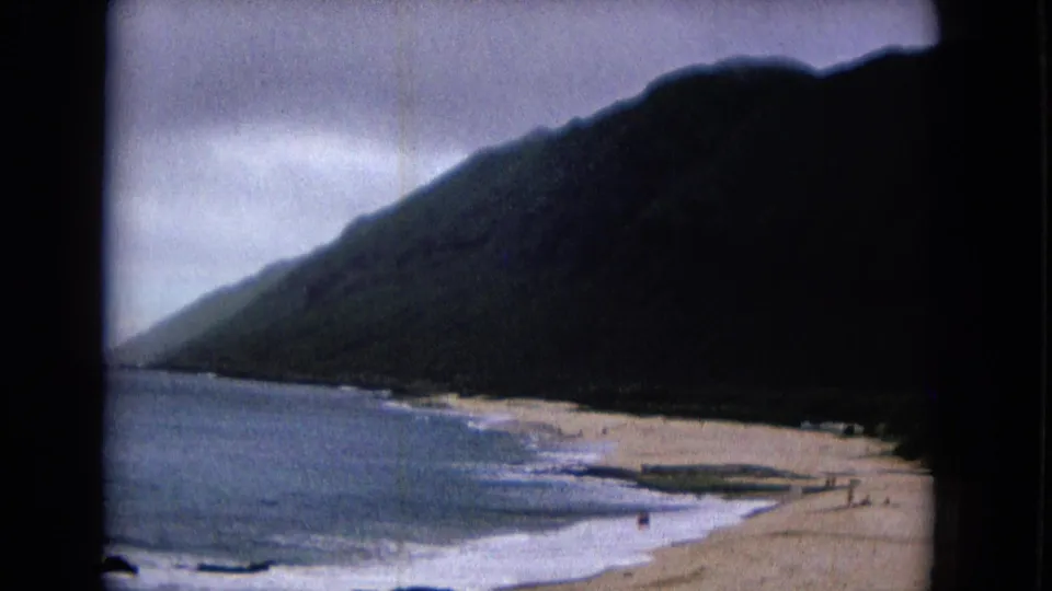 1967 8mm frame of a hillside above a sandy beach, surf, cloudy sky, and tiny distant people near the shore.