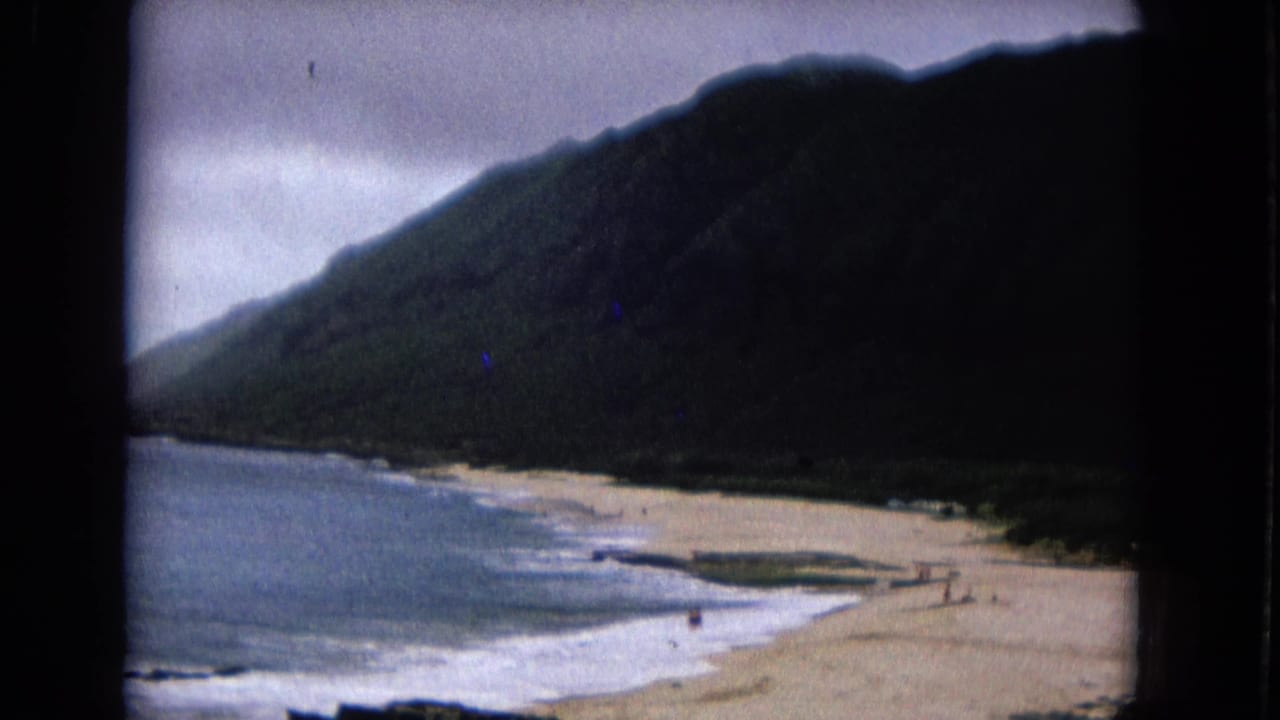 1967 8mm home-movie frame showing a sandy shoreline, surf, and dark mountain slope with distant beachgoers.