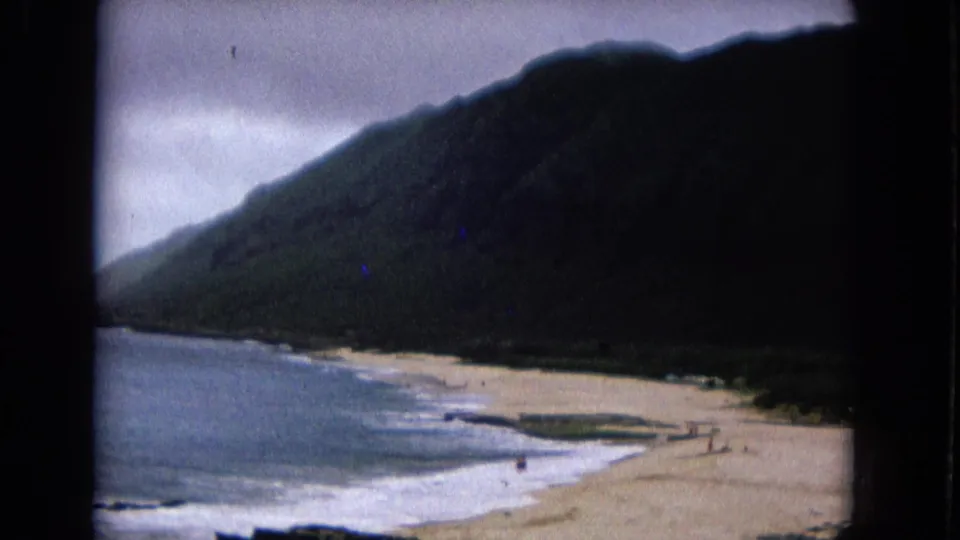 1967 8mm home-movie frame showing a sandy shoreline, surf, and dark mountain slope with distant beachgoers.