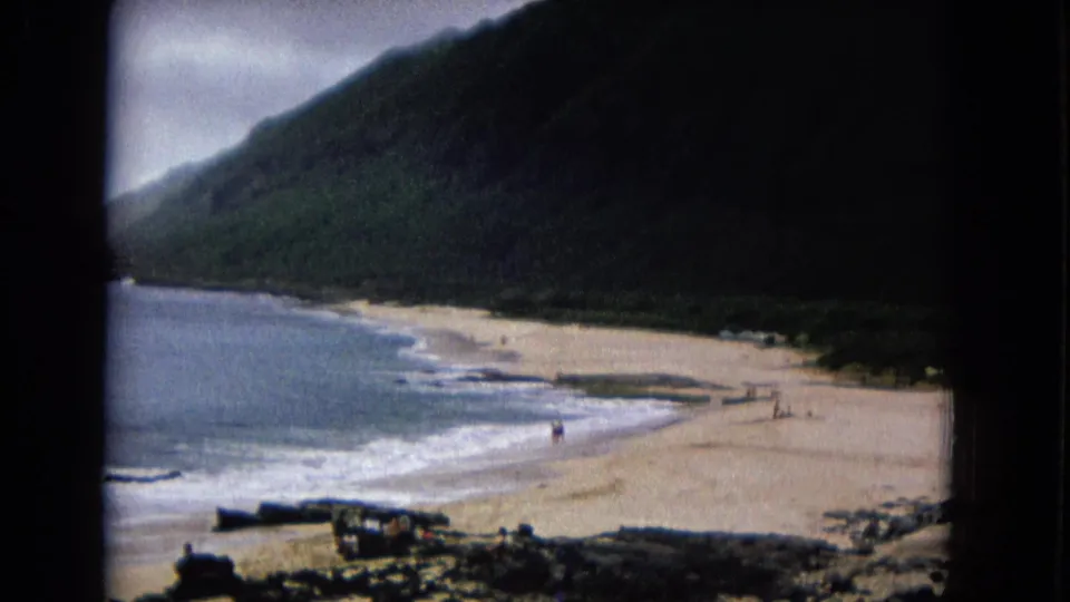 1967 home-movie frame showing surf along a sandy beach, with a shadowed mountain and driftwood or rocks in the foreground.