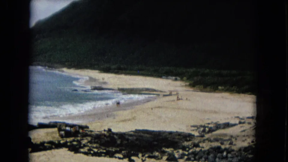 1967 8mm frame of a broad sandy beach, small beachgoers, surf, and a dark forested mountain under cloud cover.