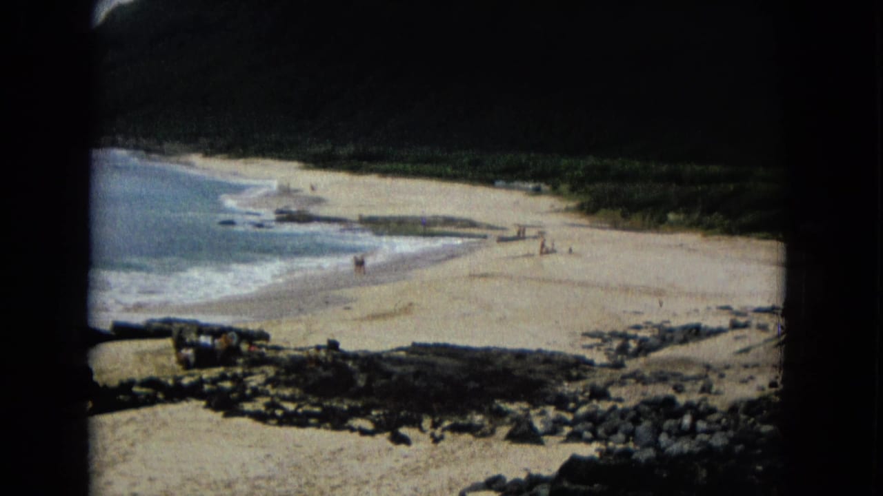 1960s 8mm frame of a sandy coastal beach with small distant people, waves, rocks, and dark green land beyond.