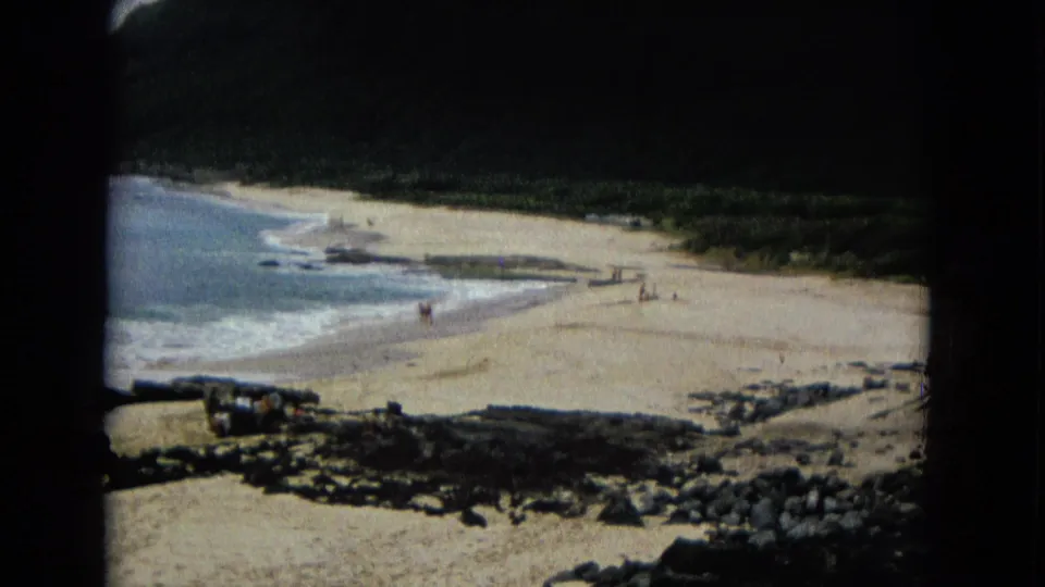 1960s 8mm frame of a sandy coastal beach with small distant people, waves, rocks, and dark green land beyond.