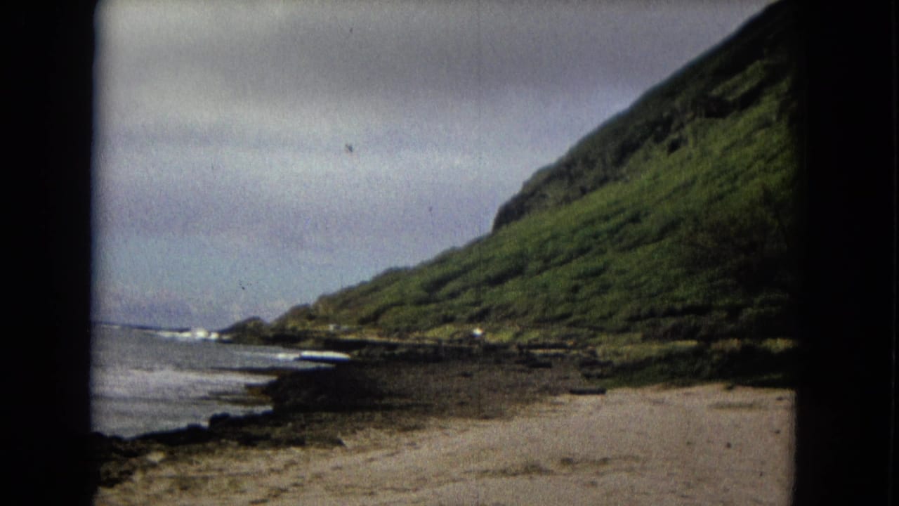 1960s 8mm coastal view of waves meeting a rocky shore beneath a steep green mountain slope.