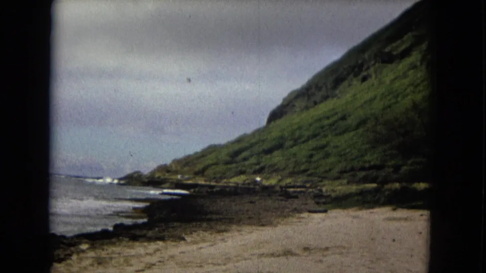 1960s 8mm coastal view of waves meeting a rocky shore beneath a steep green mountain slope.