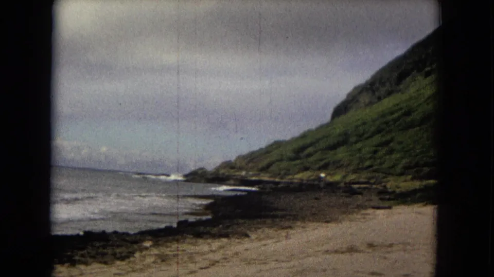 Soft 8mm frame showing surf along a dark rocky coast with a green slope descending toward the water.